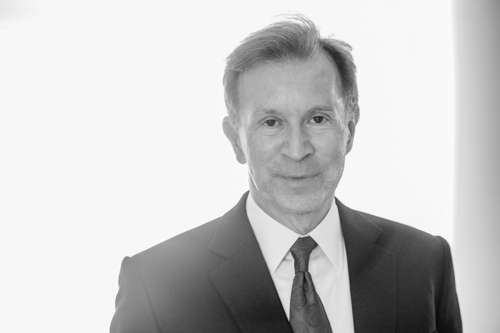 A black and white headshot of John Studzinski. He is wearing a white shirt and black suit jacket and tie, and is standing in front of a plain white background. He has a neutral expression on his face and is looking directly at the camera.