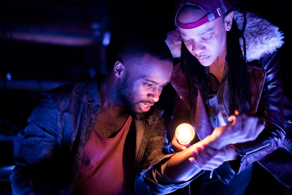 A photo of two actors from Tangle Theatre Company. One of the actors is shining a light on the other actor's arm as they hold it up in the light. They are both inspecting the arm.