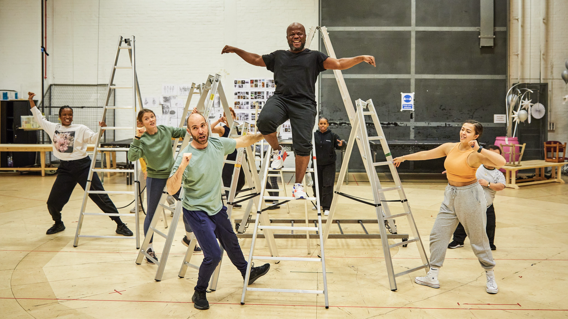 Inside a large rehearsal room with white brick walls and a wooden floor, a group of four actors stand on 3 different step ladders, smiling and singing with another actor who is stood at the top of one of the step ladders, with their arms spread out wide, laughing with a big open smile.