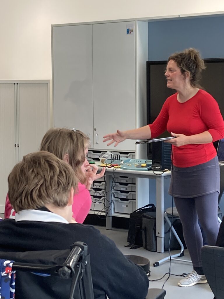 In a large classroom, a woman with curly brown hair tied up, wearing a red top and grey leggings is stood up conducting a group of students who are sat down.