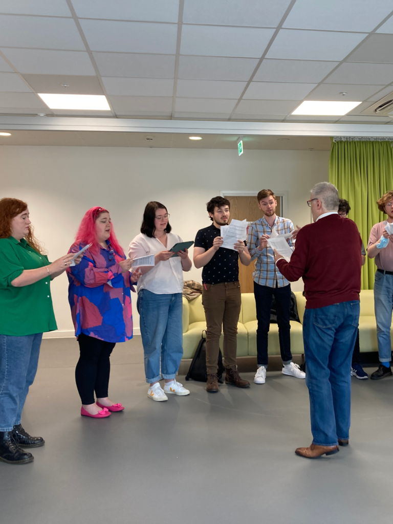 In a large classroom, seven people stand in a line holding open songbooks. James Macmillan is stood in front of them, conducting, facing away from the camera. James has short grey hair, glasses, and is wearing a burgundy jumper with a white shirt underneath. 