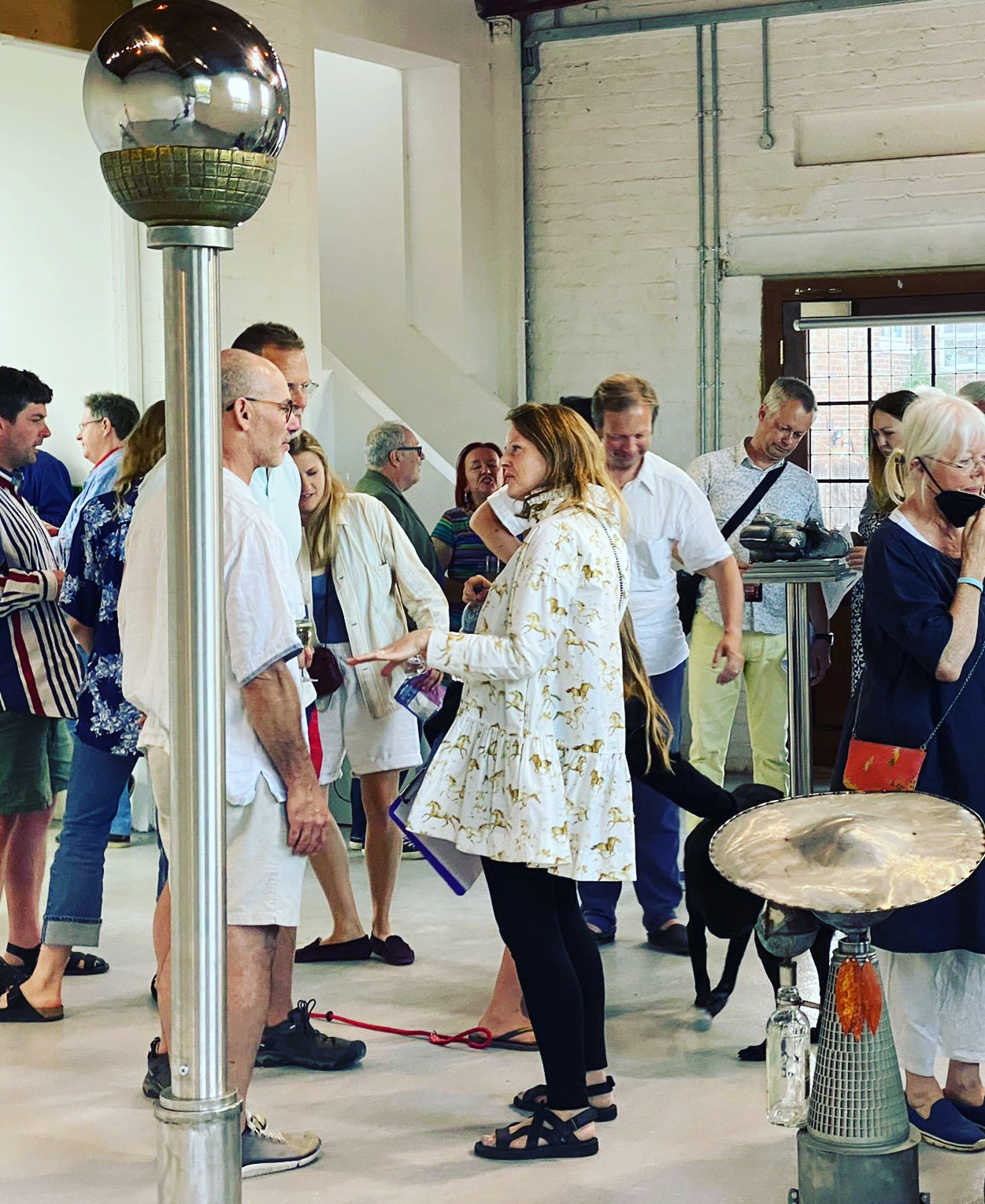 A group of people inside an art studio, looking at pieces in an exhibition. In the foreground of the photo is a tall metal sculpture with a glass orb at the top. 