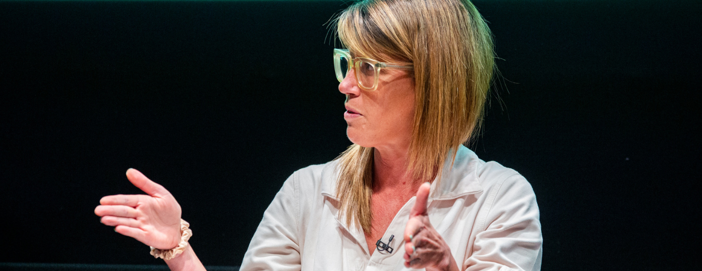 Pip Jamieson sat on a stage talking to someone off camera. She has long blonde hair and is wearing glasses with thick frames and a cream shirt.