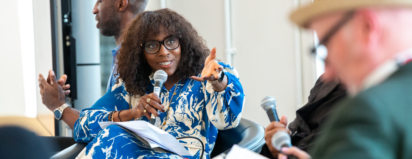 Brenda Emmanus sat on an arm chair, holding a microphone with one hand and gesticulating with her other hand.