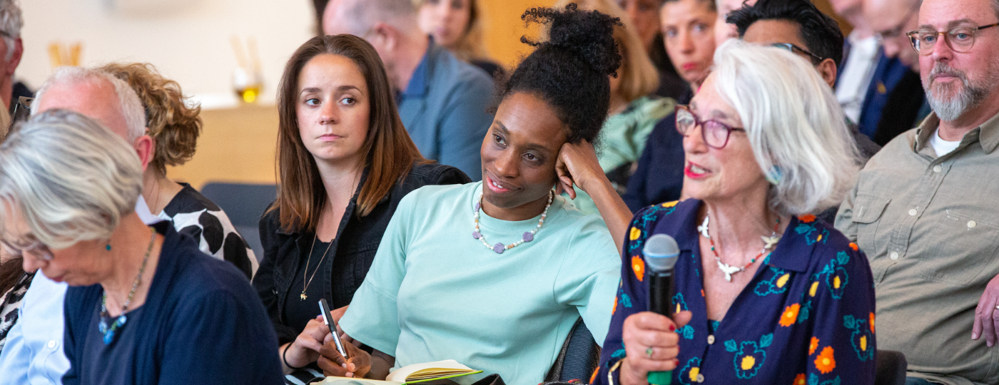 A row of people sat amongst a busy seated audience. The person furthers right is holding a microphone and speaking out. The person beside them is leaning their head on their hand, smiling, and writing in a notebook.