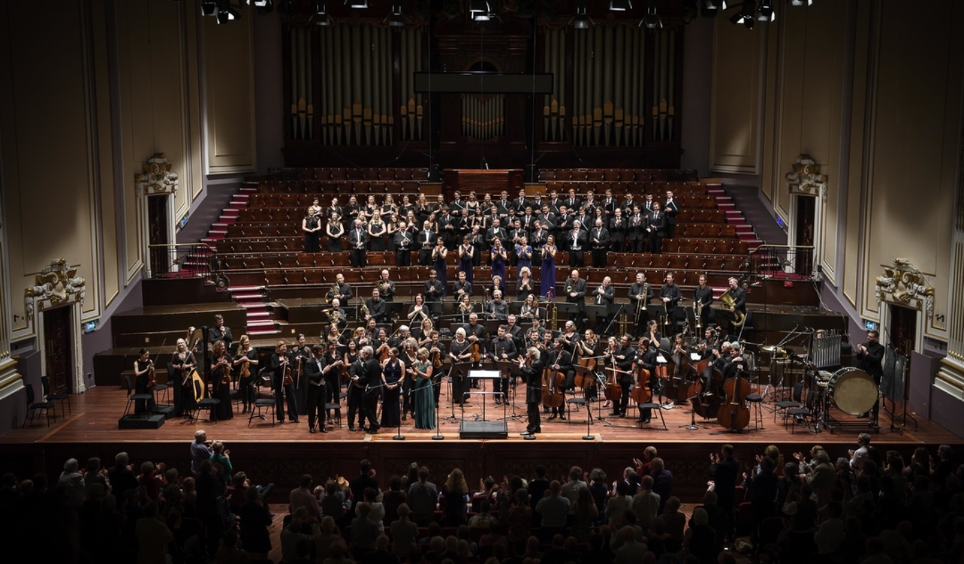 A wide shot of The Sixteen arranged in rows onstage at the Edinburgh’s Usher Hall. In front of them is a seated orchestra. In front of that is a seated audience.