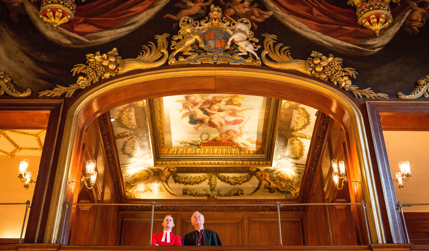 An upward angle shot of the ceiling of The Chapel Royal at Hampton Court Palace. There are paintings of royal red curtains hanging, as well as a large golden crest with a lion and unicorn on.