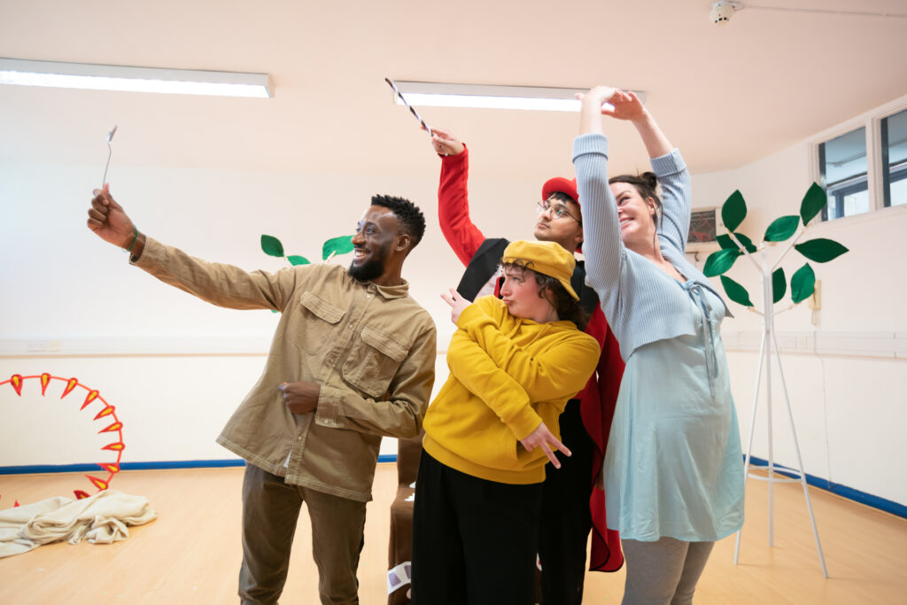 Inside a rehearsal room with coat stands propped up to look like trees, a group of four actors stand in a group, posing for a photo. The actor at the front of the group is holding up a silver spoon, which the others look into and pose, as if the spoon is a phone camera.