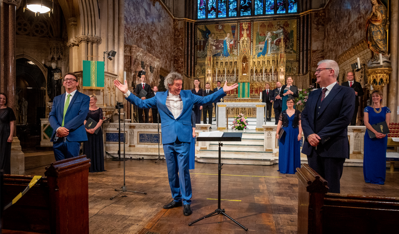 Inside Farm Street Church. It has grand stone arches and golden statues spread across its walls. Harry Christophers stands in front of members of The Sixteen choir, with his arms held up, smiling.