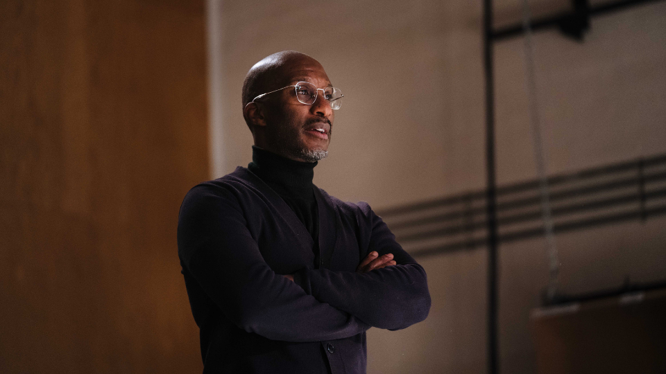 A headshot of Clint Dyer inside a rehearsal room with brown and white walls. Clint is middle aged man with a short dark grey beard. He is wearing a black high neck top and glasses, and has his arms folded in front of him. He is looking to the side, off camera with a neutral expression.