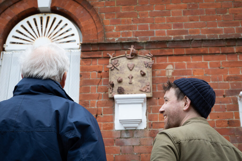 A photo of two people standing in front of a sculpture on the side of a brick wall.