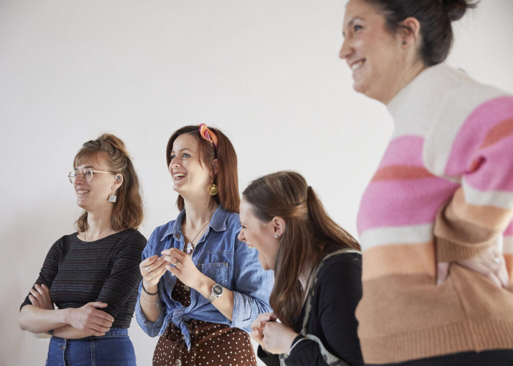 A group of women stood in front of a white wall, laughing with someone off camera.