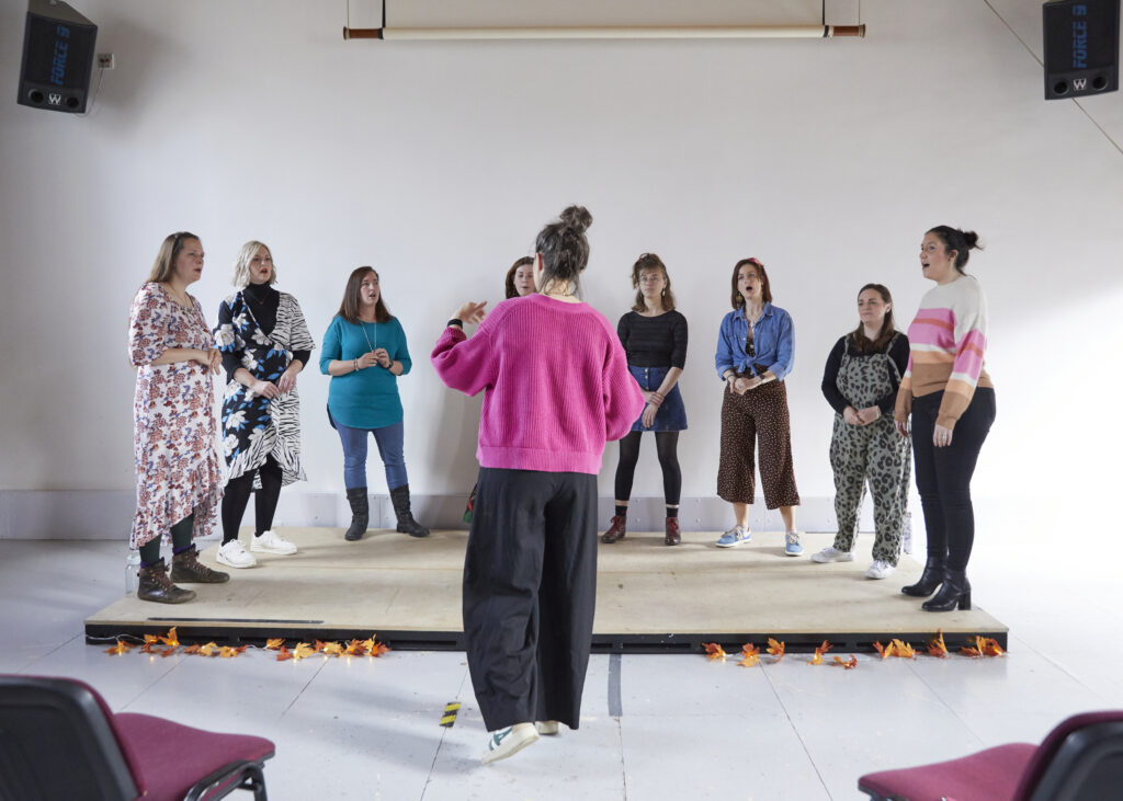 A group of women standing on a stage singing in a white room. A woman in a pink jumper stands in front of them and conducts. 