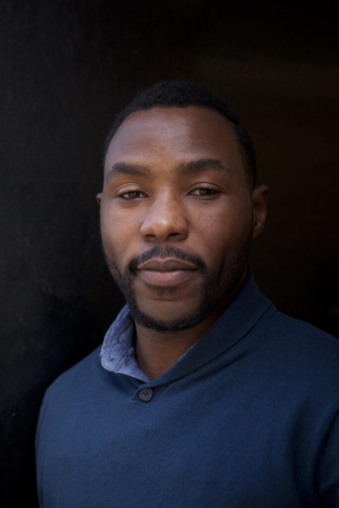 A headshot of director Gbolahan Obisesan. Gbolahan has short black hair and is wearing a smart blue jumper with a lighter blue collared shirt underneath. They are stood in front of a plain black background with a neutral expression.