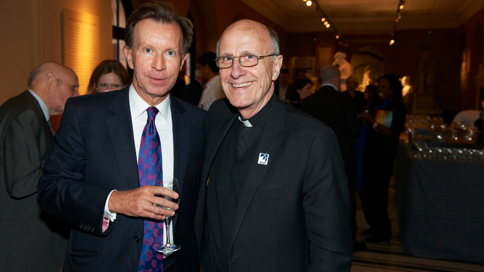 A photo of John Studzinski and Msgr Vladimir Felzmann, standing in a busy room, smiling at the camera.