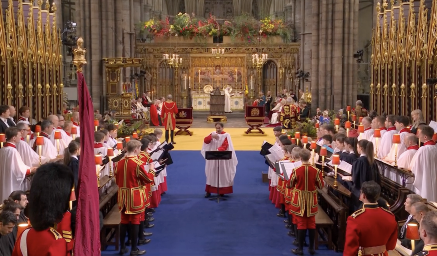 A wide shot of several rows of choral singers inside Westminster Abbey. The rows of singers are facing each other, holding scores and singing out. In the centre at one end of the singers is Andre Nethsinga, who is conducting.