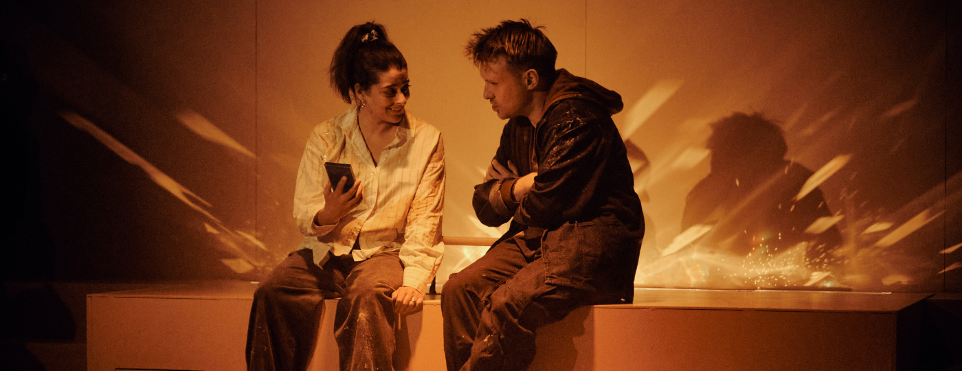 A small, dark haired woman with mid length hair in a ponytail and a man with sandy hair and blue eyes sit on a cabinet built into an orange wall. They are both covered in custard. The woman holds up a small block-shaped object to show to the man while he speaks.