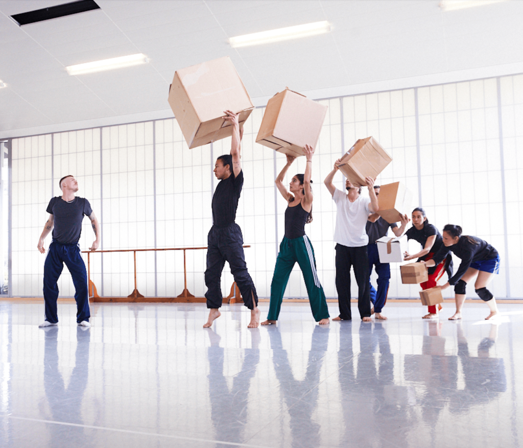 A photo of members of Akram Khan Company rehearsing in a bright studio. The dancers are in a line each holding a cardboard box above their heads. One dancer stands alone looking up at the boxes.