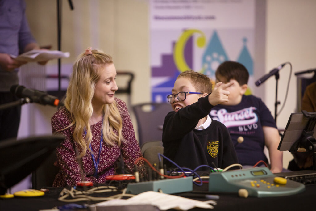 An adult and a child in schoolk uniform sat at a desk with cables and sound monitors on. The child is pointing at something off camera and smiling at the adult. The adult is smiling back at them.